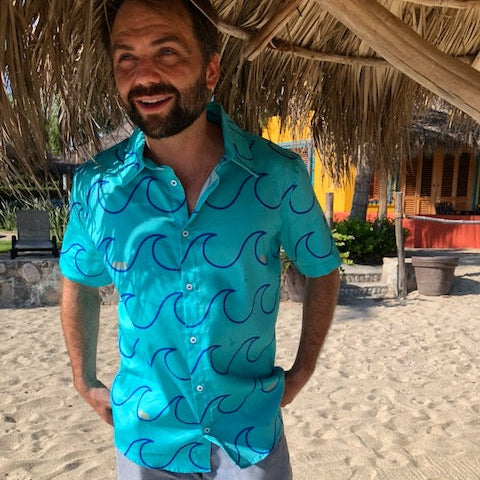 A man standing on a beach wearing a blue short-sleeved organic cotton Aloha shirt with a wave pattern and mother of pearl buttons, paired with light shorts.