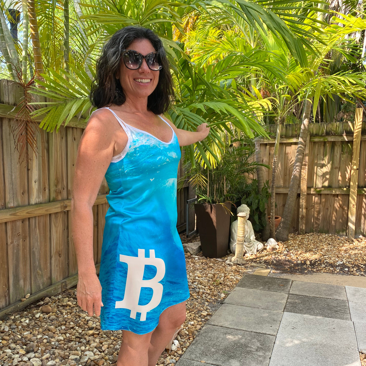 A woman wearing a bright blue tank top and matching blue shorts with a large white Bitcoin symbol, posing outdoors on a stone patio with plants and a wooden fence in the background.
