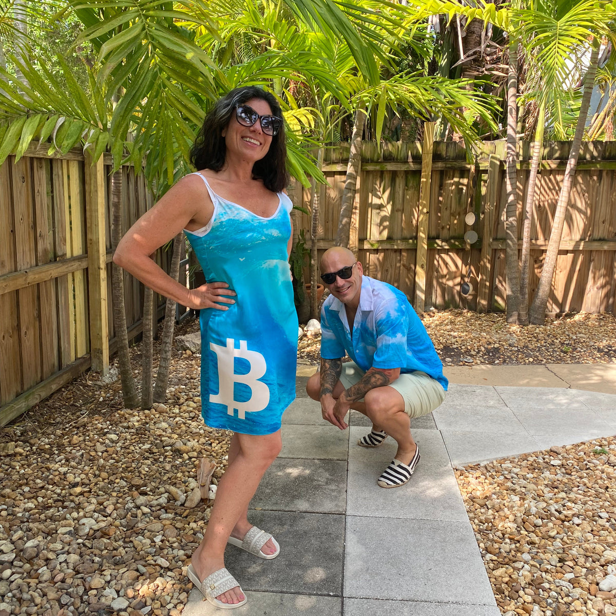 A woman wearing a bright blue sleeveless dress with a large Bitcoin symbol on the front, posing outdoors on a paved walkway with palm trees and gravel in the background. A man in a blue shirt is also visible in the scene.
