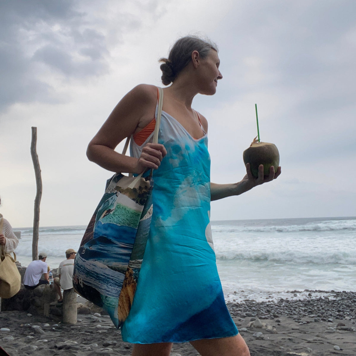 A woman on a beach wearing a blue and green patterned sarong dress, holding a coconut drink with a straw.