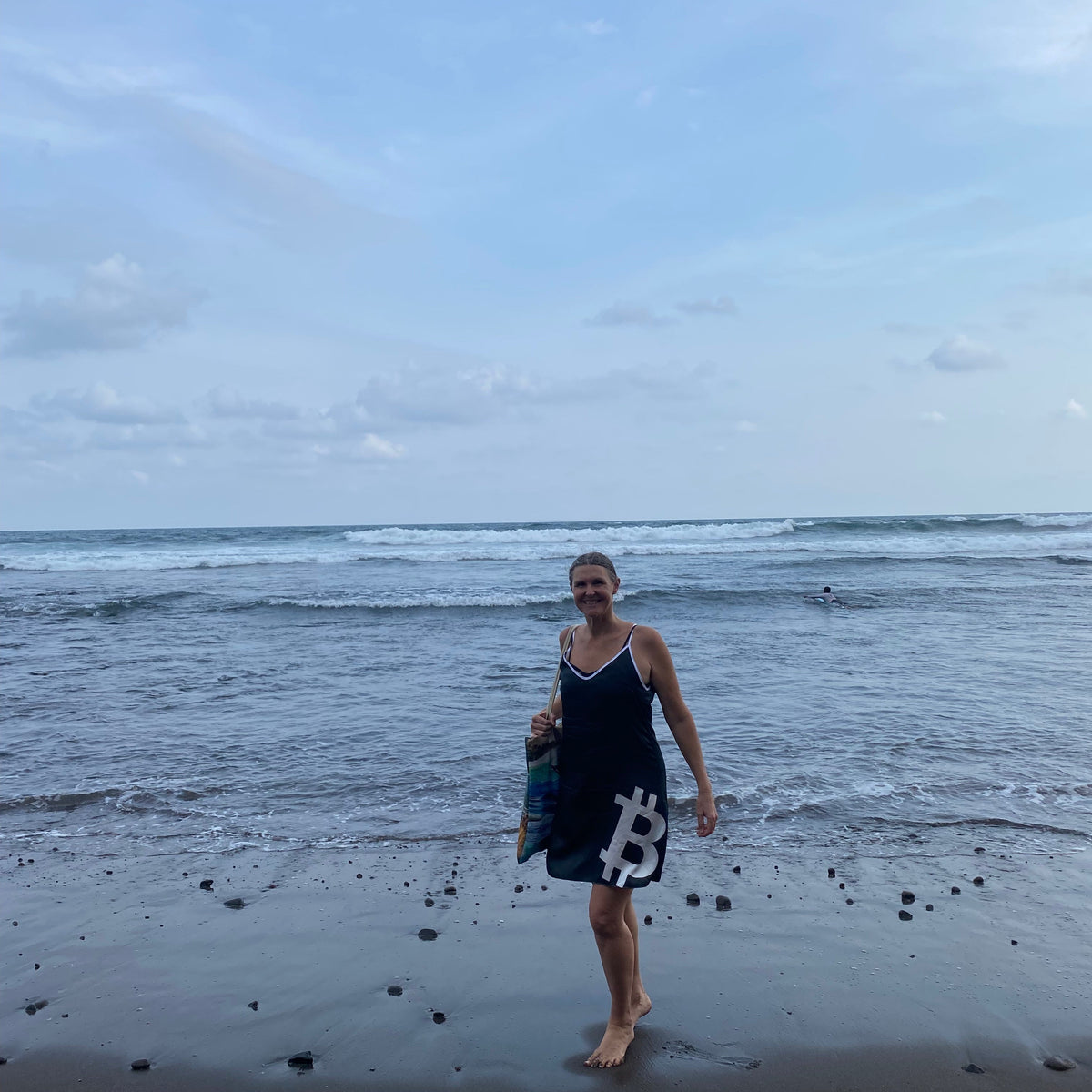 A woman walking on a sandy beach near the shoreline, wearing a dark sleeveless slipdress with a white geometric B for Bitcoin.